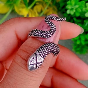 Puede incluir: Un anillo plateado con forma de serpiente que se enrolla alrededor de un dedo. El anillo presenta escamas detalladas y una cabeza de serpiente con un diseño en forma de cruz. El cuerpo de la serpiente se curva hacia arriba por el dedo.