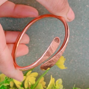 May include: A copper-colored cuff bracelet with an open design. The bracelet has a hammered texture on the ends and a smooth, rounded band. The bracelet is held by a hand, with a green and yellow floral background.