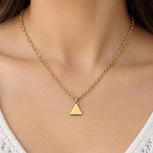 May include: A gold-coloured necklace featuring a delicate chain and a small, solid triangle pendant. The pendant hangs centrally from the chain, which is composed of uniform oval links. The metal has a smooth, polished finish throughout the piece.