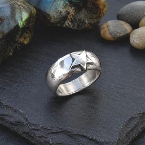 May include: A polished silver-toned ring featuring a single, raised five-pointed star emblem on its outer band. The ring has a smooth, rounded surface and a reflective metallic finish, showcasing a clean, simple design with a prominent star detail.