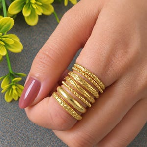 May include: A set of six gold-toned rings stacked on a finger. The collection includes bands with varied textures, including smooth surfaces, beaded edges, and intricate engraved patterns, all featuring a consistent metallic yellow finish.