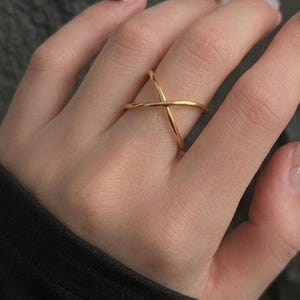 May include: A polished gold-tone ring featuring a sleek, crossover design where two thin, smooth bands intersect to form an X shape. The ring has a continuous, rounded profile and a reflective metallic finish that catches the light across its surface.
