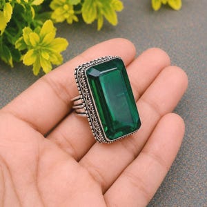 May include: A rectangular emerald green gemstone ring set in a silver band with intricate beaded and scrollwork detailing along the edges. The large, faceted stone is positioned vertically, showcasing a deep green hue and a polished finish.