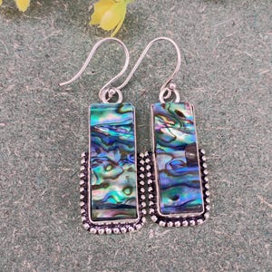 May include: A pair of silver-tone earrings with rectangular abalone shell pendants. The iridescent pendants display shades of blue, green, and purple. The edges of the pendants are framed with a beaded silver border and have a hook closure.
