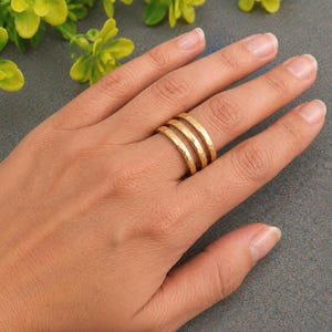 Può includere: Un anello color oro caratterizzato da tre sottili fasce sovrapposte con una finitura martellata e testurizzata. L'anello è indossato su un dito, mettendo in risalto il design a strati delle fasce metalliche unite in un unico gioiello.