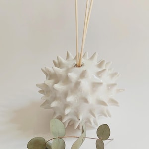 Peut inclure: Un diffuseur en céramique blanche avec un design sphérique et épineux. Le diffuseur contient plusieurs bâtonnets en bois et est décoré d'une branche de feuilles vertes. Produit pour l'aromathérapie.