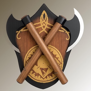 May include: A decorative wall hanging featuring two crossed axes. The axes have black blades and wooden handles, set against a wooden shield with gold-colored Celtic knot and emblem designs. The shield has a black border.
