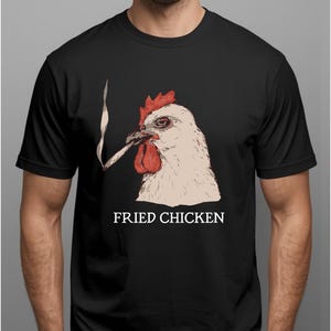 Peut inclure: T-shirt noir avec un graphisme représentant un poulet fumant une cigarette, avec les mots "FRIED CHICKEN" en lettres blanches. Le poulet est blanc avec une crête et des barbillons rouges.