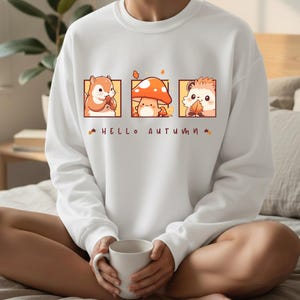 May include: White sweatshirt featuring three autumn-themed illustrations: a squirrel, a mushroom, and a hedgehog. The phrase "HELLO AUTUMN" is printed below the illustrations. The sweatshirt is made of a soft material.