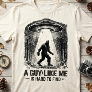 Puede incluir: Camiseta color crema con un gráfico de Bigfoot siendo abducido por un OVNI en un entorno forestal. El texto dice "A Guy Like Me Is Hard to Find" en negro. El diseño es una versión humorística del críptido.