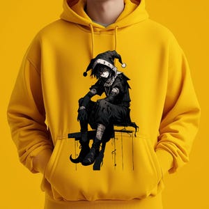 May include: Mustard yellow hoodie featuring a black and white graphic of a seated anime character with a jester hat. The character is wearing black clothing and boots. The hoodie has a drawstring hood and a front pocket.