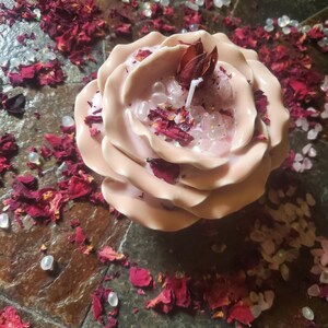 Könnte beinhalten: Eine hellrosa rosenförmige Kerze mit einem weißen Docht, verziert mit getrockneten Rosenblättern und kleinen Kristallen. Die Kerze ist von verstreuten Rosenblättern und kleinen, klaren Perlen umgeben, was eine romantische und dekorative Ästhetik schafft.