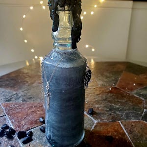 Peut inclure: Une bouteille en verre transparent remplie d'un liquide sombre et tourbillonnant. La bouteille est scellée avec de la cire noire et ornée d'une chaîne et de pendentifs argentés. Des pierres noires sont éparpillées à la base, créant une esthétique gothique.