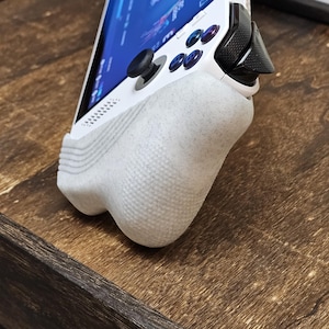 May include: A white handheld gaming device with a screen displaying a game interface. The device has a textured grip and black control sticks and buttons. The device is resting on a wooden surface.