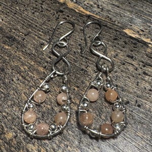 May include: A pair of handcrafted earrings featuring a teardrop design. The earrings are made with silver-colored wire and adorned with small, round, peach-colored beads. The earrings have a hook closure.