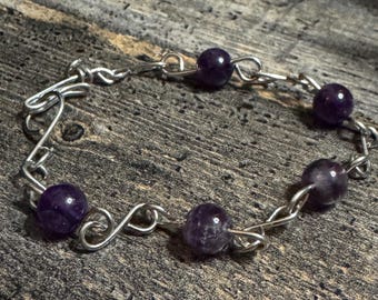May include: A silver-toned bracelet featuring six round, deep purple amethyst beads. The beads are connected by a chain-like design with looped links. The bracelet is displayed on a weathered, gray wooden surface.