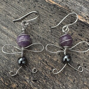May include: A pair of handmade earrings featuring a purple amethyst bead and a dark gray bead, connected by silver wire. The earrings have a unique design with looped wire wings and a hook closure. The earrings are displayed on a wooden surface.