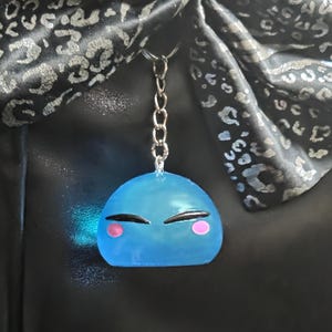Peut inclure: Un porte-clés bleu en forme de slime avec une chaîne argentée. Le slime a les yeux noirs fermés et des joues roses. L'arrière-plan comprend une surface noire et un tissu noir et argenté.