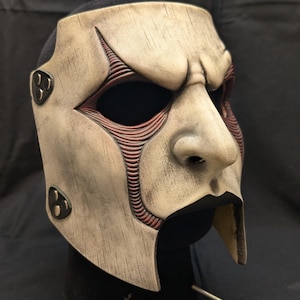 May include: A weathered, off-white mask with a prominent nose and deep eye sockets. Reddish-brown lines detail the cheek and eye areas. The mask has a distressed finish and is mounted on a black stand, suggesting a theatrical or costume piece.