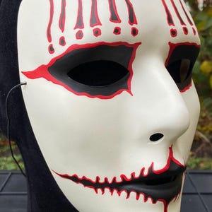 May include: A white mask with black eye holes and a black mouth, outlined in red. Red exclamation marks are painted above the eyes. The mask is on a black head form, with a black string attached.