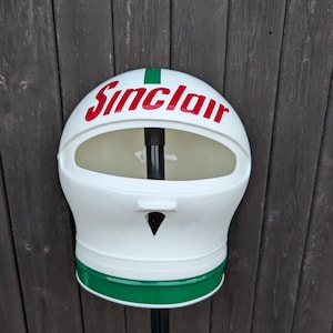 May include: A white and green Sinclair gas station trash can. The trash can has a rounded top with the word "Sinclair" in red lettering. A green stripe accents the top and bottom of the can. The trash can is mounted on a black pole.