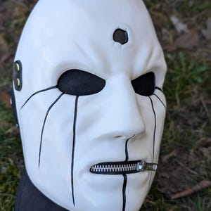 May include: A white mask with black accents, featuring a zipper mouth and black tear-like lines. The mask has a small hole above the right eye and black eye sockets. The mask is set against a blurred green and brown background.