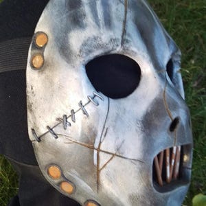 May include: A distressed, metallic-looking mask with a stitched design and visible teeth. The mask has a weathered appearance with gray and white paint, and is secured with a black strap. The mask has a menacing look.