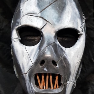 May include: A metallic silver mask with a distressed, cracked appearance. The mask features large eye openings, a nose, and an open mouth revealing wooden slats. A leather strap is attached at the top.