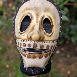 May include: A distressed, off-white mask with a zipper mouth and large eye openings. The mask features a prominent nose, stitched details, and is adorned with brass-colored rivets. The top and bottom edges are black.