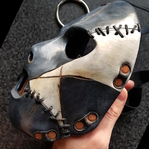 May include: A handcrafted, multi-toned leather mask with a dog-like design. The mask features a combination of gray, white, and black leather, stitched together with black thread. It has a metal ring at the top and is held by a black strap.