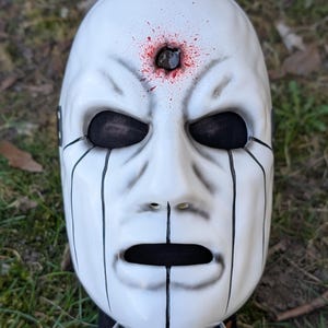 May include: White mask with black eye and mouth openings, and black tear-like lines. The mask has a hole in the forehead with red splatter details. The mask is held up by a metal stand.