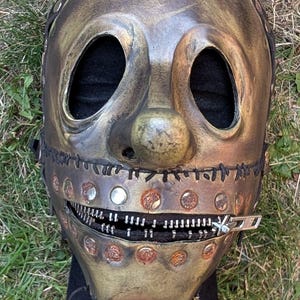 May include: A distressed, metallic-toned mask with large eye openings and a prominent nose. The mask features a zipper mouth and is adorned with rivets and stitching. The mask is designed to be worn and is placed on a black base.