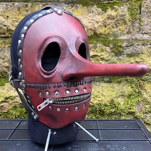 May include: A red and black leather mask with a long nose and a zipper mouth. The mask has large eye holes and is adorned with metal studs and straps. The mask is designed to be worn and is likely for costume or theatrical purposes.