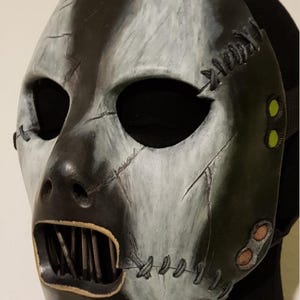 May include: A two-tone mask with a distressed, industrial aesthetic. The mask is split vertically, with one side painted light gray and the other black. It features stitched details, eye holes, and a mouth with metal bars.