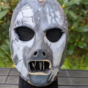 May include: A distressed, gray and white mask with visible stitching and cracks. The mask has a leather strap and rivets at the top. The mouth area features a metallic grill. The mask is displayed on a black stand.