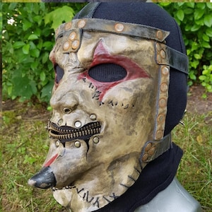 May include: A distressed, full-face mask with a zipper mouth and stitched details. The mask features a weathered, tan finish with red accents around the eyes and a black nose. Metal rivets and leather straps add to the rugged aesthetic.