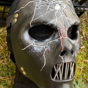 May include: Black mask with a cracked design and metal teeth. The mask has white lines that resemble cracks across the face and red accents near the nose. The mask is secured with a black strap and gold rivets.