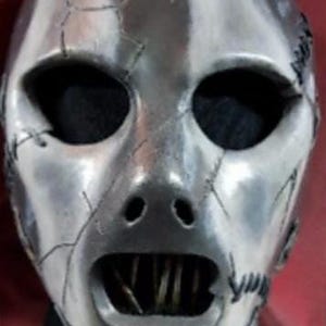 May include: A silver-colored mask with a distressed, cracked appearance. The mask has large, oval eye openings and a wide mouth revealing a row of teeth. The mask's design suggests a horror or theatrical theme.