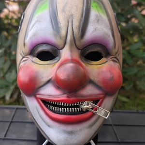 May include: A clown mask with a zipper mouth. The mask has a pale complexion with red cheeks and nose, purple eye shadow, and green accents above the eyes. The zipper is silver.