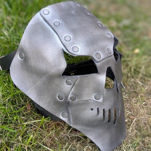 May include: A gray leather mask with a skull-like design. The mask features riveted details and a black strap. The eye and mouth openings are visible. The mask is designed to cover the face.