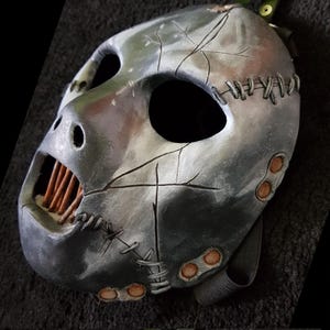 May include: A metallic gray mask with a stitched design and visible cracks. The mask has large eye openings, a wide mouth with visible teeth, and a strap for wearing. The mask is likely for costume or decorative purposes.