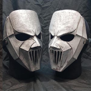 Pode incluir: Duas máscaras angulares prateadas com aberturas escuras para os olhos e fendas verticais para a boca. As máscaras têm um acabamento desgastado e texturizado, sugerindo um material metálico. São exibidas em cabeças de manequim pretas.