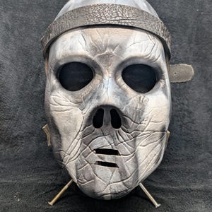 Puede incluir: Una máscara gris y blanca con un aspecto envejecido, con grandes aberturas para los ojos y una superficie texturizada detallada. La máscara tiene una correa de cuero alrededor de la cabeza y está montada en un soporte metálico. El diseño evoca una sensación de edad y misterio.