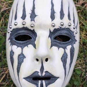 May include: A white mask with black accents and silver studs. The mask features black tear-like streaks, eye markings, and a stylized mouth. The mask has a smooth, matte finish and is designed for costume or decorative purposes.
