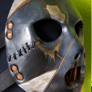 May include: A distressed black mask with a stitched design and metallic accents. The mask features a cracked appearance with brown and silver highlights. The eye holes are large and dark, and the mouth area has a visible opening.