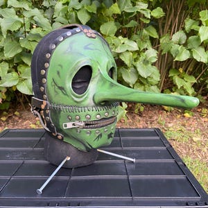 May include: A green plague doctor mask with a long, curved beak and a black hood. The mask features a zipper across the mouth and is adorned with metal studs and straps. The mask is displayed on a black surface against a backdrop of green foliage.