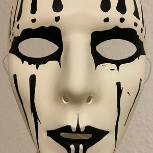 May include: A white mask with black accents. The mask features black markings around the eyes, mouth, and forehead, with vertical lines extending downwards. The mask has eye holes and a nose opening, and is likely for costume or decorative purposes.