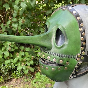 May include: A green leather plague doctor mask with a long beak, eye holes, and a zippered mouth. The mask is attached to a gray head form with black leather straps and brass rivets. The mask is a historical costume piece.