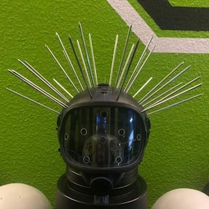 May include: A black helmet with a clear visor and numerous silver spikes protruding from the top. The helmet has a round shape and is set against a green background.