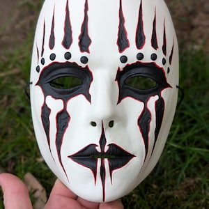 May include: A white mask with black and red accents. The mask features black eye sockets, lips, and tear-shaped designs. Black dots and lines adorn the forehead. The mask is held by a hand against a blurred green background.